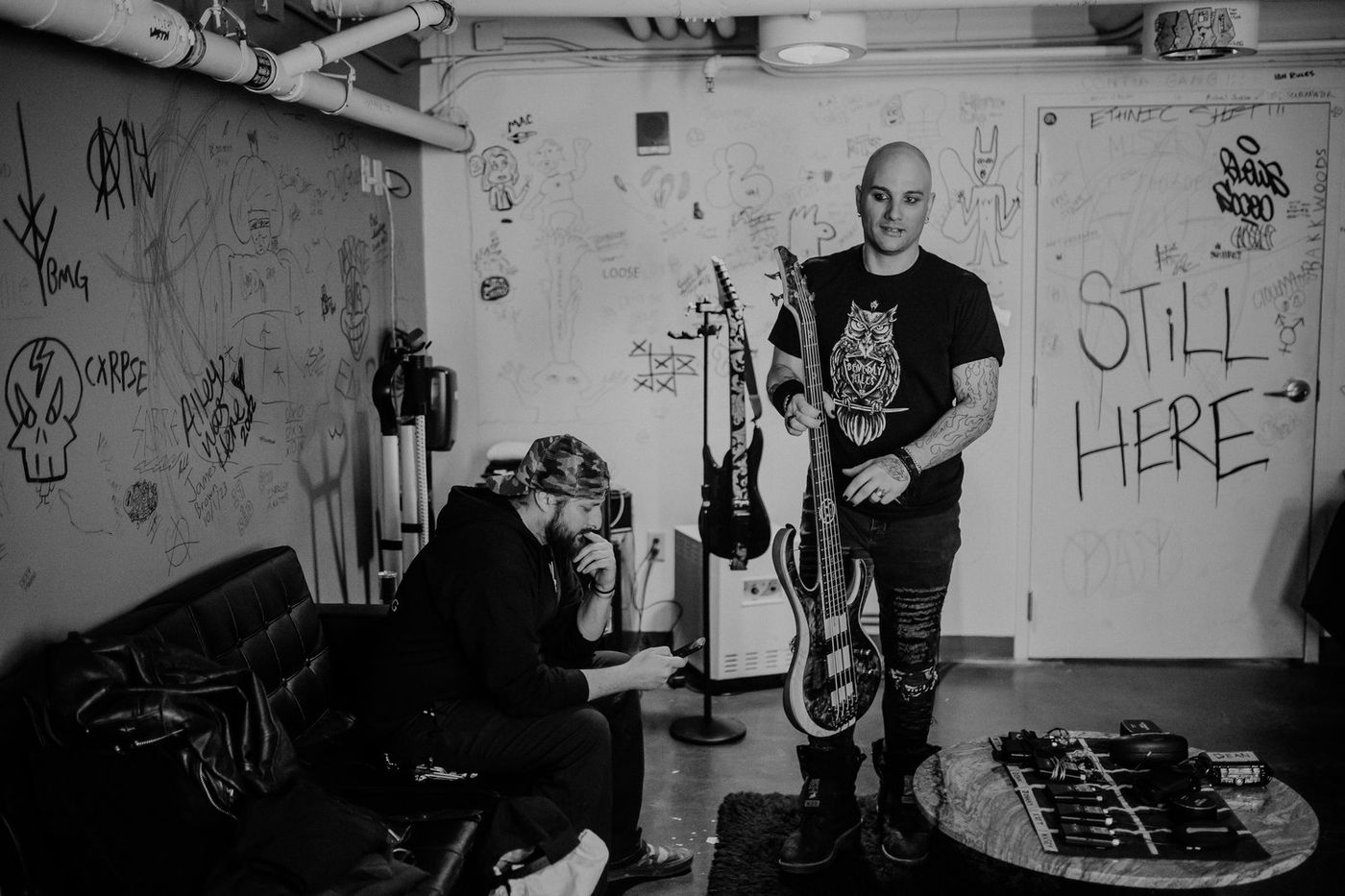 Dean in a graffitied green room holding his five-string, STILL HERE written on the door